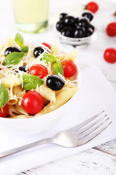 Pasta with tomatoes, olives and basil leaves in bowl on napkin on wooden background — Stock Photo, Image