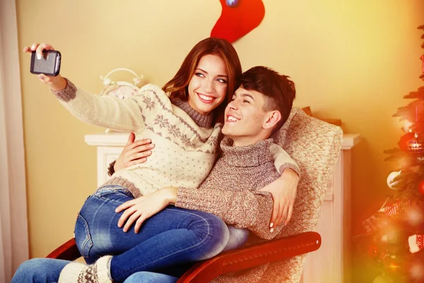 Giovane coppia con cellulare seduto sulla sedia a dondolo vicino all'albero di Natale — Foto Stock