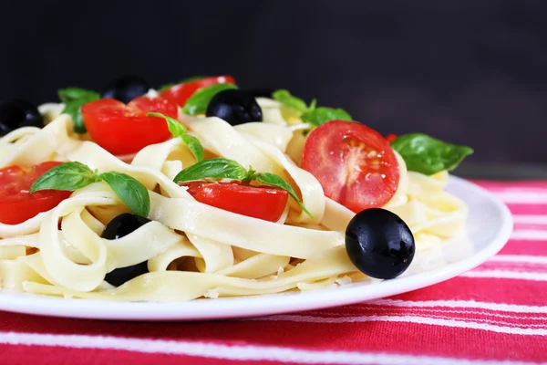 Spaghetti with tomatoes, olives and basil leaves on plate on napkin on table on wall background — Stock Photo, Image