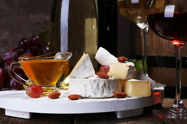 Ужин, состоящий из сыра Камамбер, меда, вина и винограда на стенде и бочки вина на деревянном столе на коричневом фоне — стоковое фото