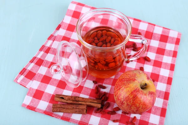 Goji berries drink in glass cup, ripe apple and cinnamon on red checkered napkin on light blue wooden background — Stock Photo, Image