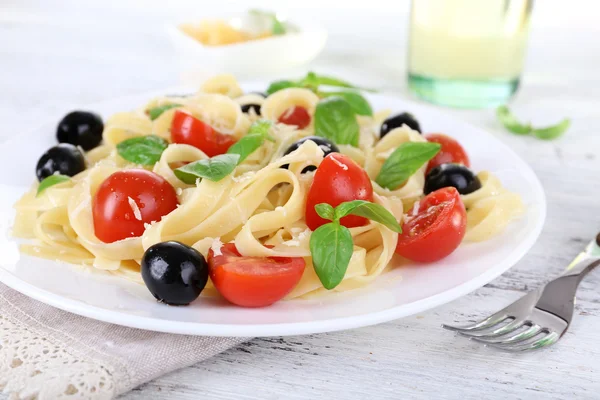 Spaghetti with tomatoes, olives and basil leaves on plate on napkin on wooden background — Stock Photo, Image