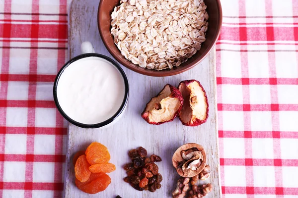 Bowl of oatmeal, mug of yogurt, marmalade, chocolate, raisins, dried apricots and walnuts on wooden cutting board on checkered fabric background