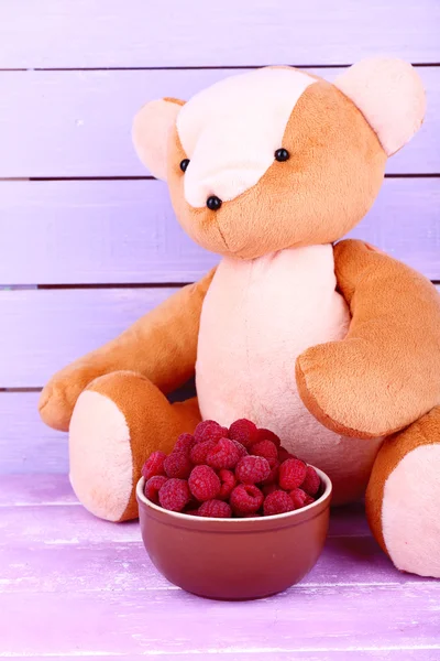 Toy bear and bowl of raspberries on wooden table on wooden wall background — Stock Photo, Image