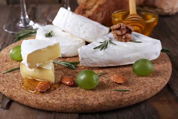 Camembert cheese on paper, grapes, nuts and honey in glass bowl on on cutting board on wooden background — Stock Photo, Image