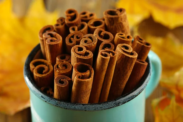 Cinnamon sticks in mug with yellow leaves on wooden background — Stock Photo, Image