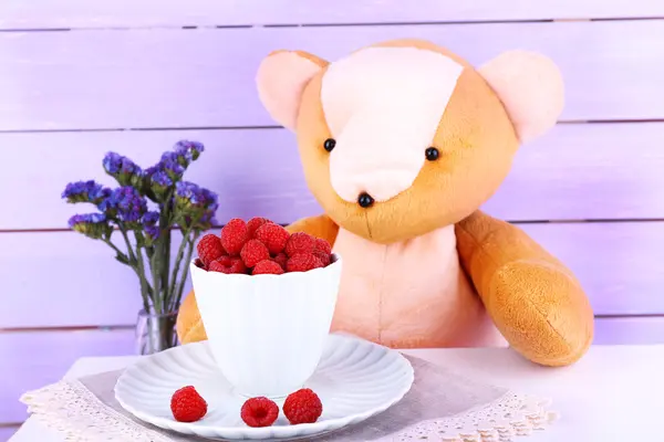 Oso de juguete con flores y taza de frambuesas sobre fondo de pared de madera —  Fotos de Stock