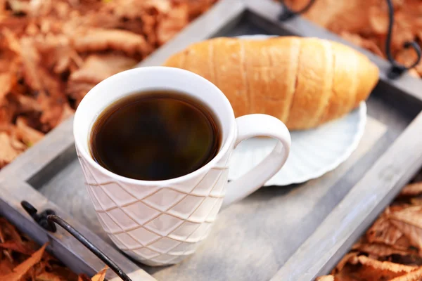 Beker van lekkere warme drank en verse croissants op dienblad, op herfst bladeren achtergrond — Stockfoto