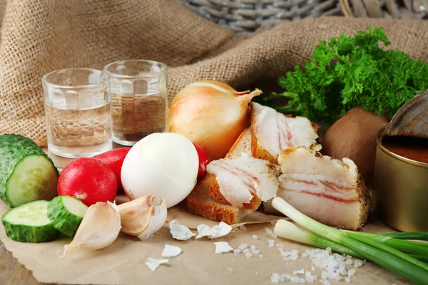 Speck, frisches Gemüse, gekochtes Ei und Brot auf Papier, Gläser mit Wodka auf Holzgrund. Dorffrühstückskonzept. — Stockfoto
