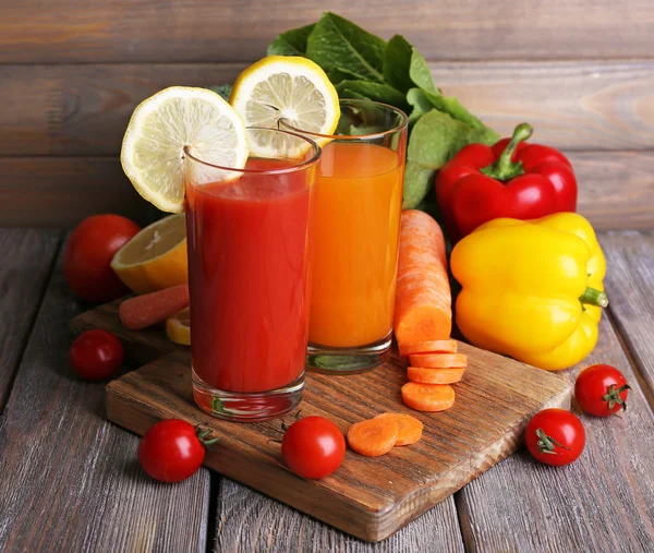 Zanahoria y jugo de tomate en vasos y verduras frescas sobre tabla de cortar sobre mesa de madera sobre fondo de pared de madera —  Fotos de Stock