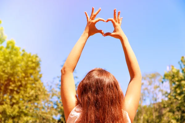 Молодая девушка держит руки в форме сердца, обрамляясь на фоне голубого неба — стоковое фото