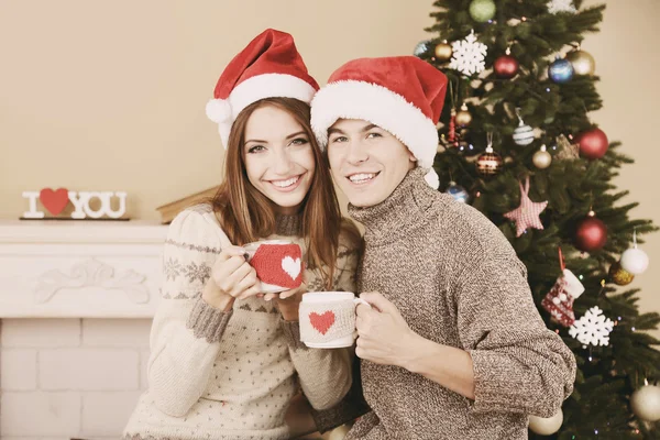 Bella coppia d'amore seduta con tazze davanti al camino vicino all'albero di Natale. Donna e uomo che celebrano il Natale — Foto Stock