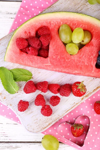 Fresh juicy watermelon slice  with cut out heart shape, filled fresh berries, on cutting board, on wooden background — Stock Photo, Image