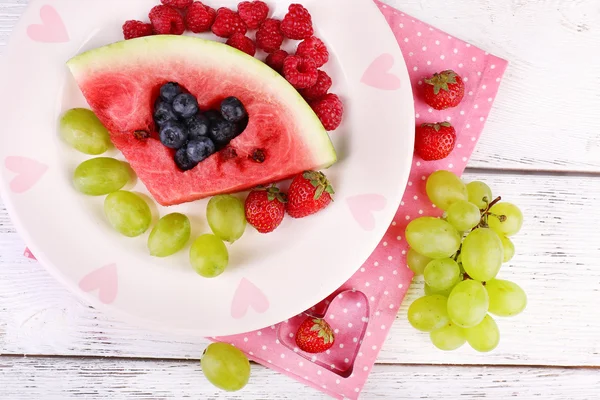 Fetta di anguria fresca succosa con forma a cuore ritagliato, bacche fresche ripiene, su piatto, su sfondo di legno — Foto Stock