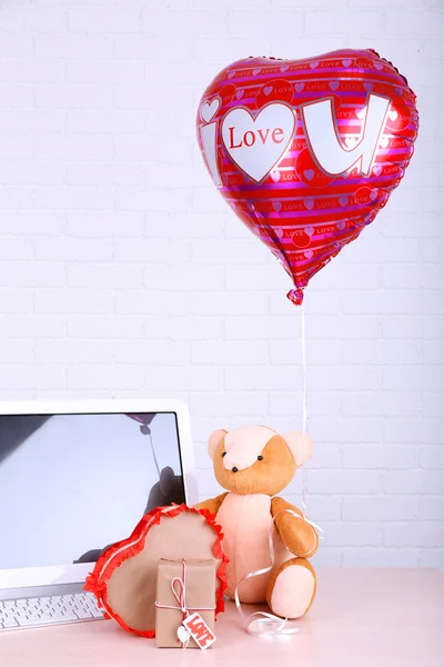 Osito de peluche con cajas presentes y globo de amor en la mesa de madera de la computadora, en el fondo de la pared de ladrillo — Foto de Stock