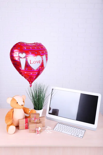 Teddybär mit Geschenkschachteln, Pflanzen- und Liebesherz-Ballon auf hölzernem Computertisch, auf Backsteinmauer-Hintergrund — Stockfoto