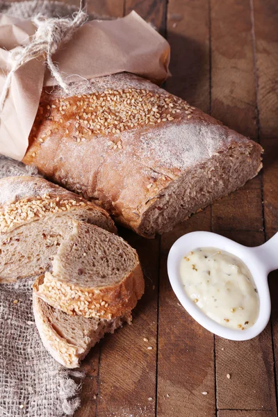 Fresh baked bread wrapped in paper and bowl with sauce, on wooden background — Stock Photo, Image