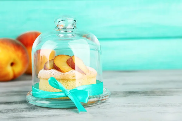 Mini gâteau savoureux avec pêche fraîche sur une soucoupe en verre avec couvercle, sur une table en bois — Photo