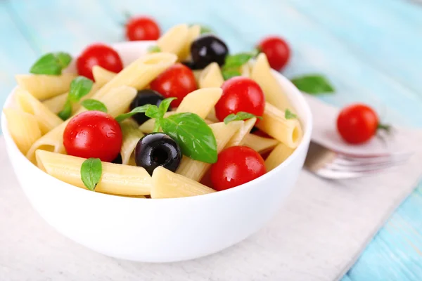 Pasta with tomatoes, olives and basil leaves in bowl on napkin on wooden background — Stock Photo, Image