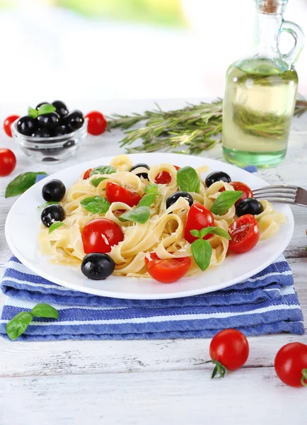 Spaghetti with tomatoes, olives and basil leaves on plate on napkin on wooden background — Stock Photo, Image