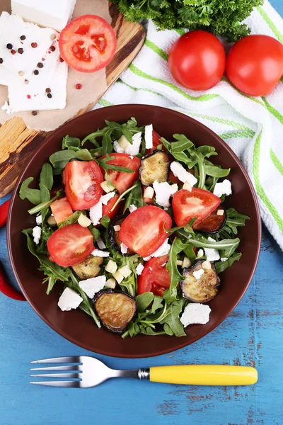 Eggplant salad with tomatoes, arugula and feta cheese, on napkin, on color wooden background — Stock Photo, Image