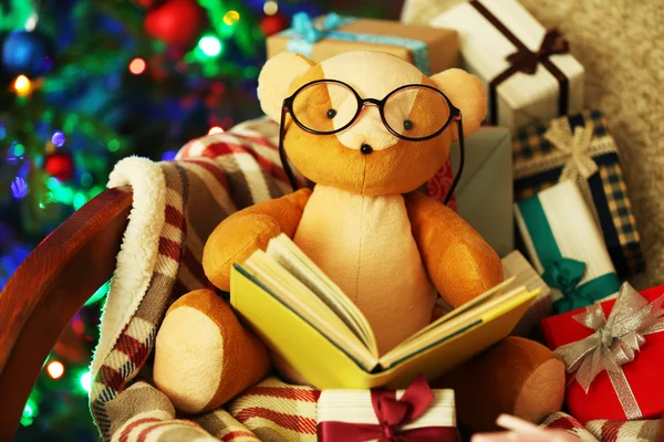 Teddy bear with book and gift boxes in rocking chair on Christmas tree background