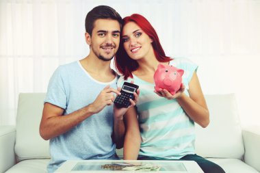 Loving couple sitting in sofa with piggy bank in room clipart