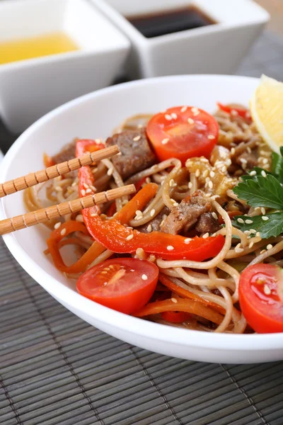 Chinese noodles with vegetables and roasted meat in bowl on napkin, on wooden background — Stock Photo, Image