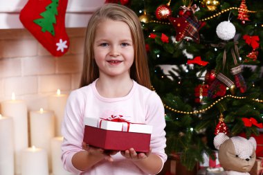 Little girl holding present box near Christmas tree on fireplace with candles background