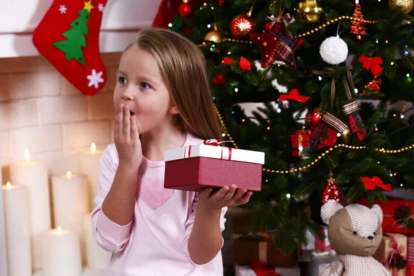 Little girl holding present box near Christmas tree on fireplace with candles background — Stok fotoğraf