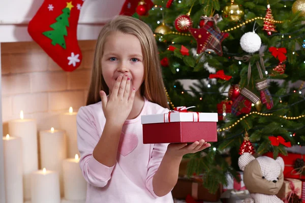 Little girl holding present box near Christmas tree on fireplace with candles background — Stockfoto