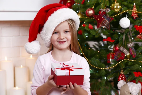 Little girl in Santa hat holding present box near Christmas tree on light background — Stock Photo, Image