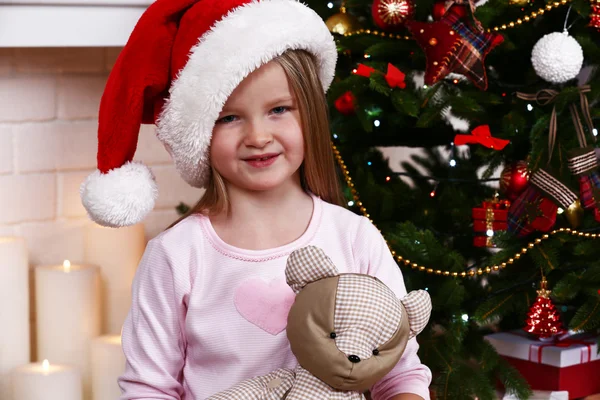 Niña en Santa sombrero con oso de peluche cerca de abeto en la chimenea con velas de fondo —  Fotos de Stock