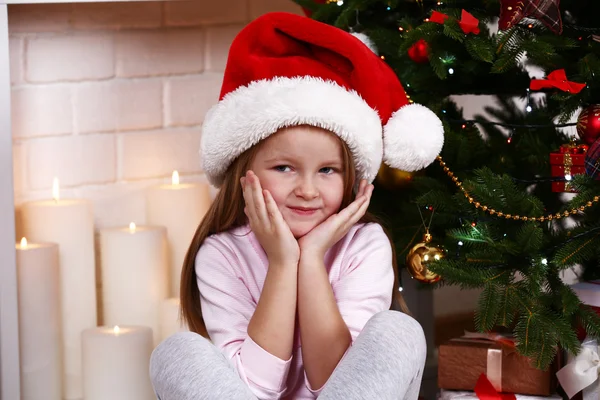 Little girl in Santa hat sitting near fir tree on fireplace with candles background — ストック写真