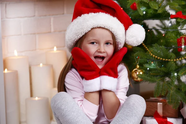 Petite fille dans le chapeau de Père Noël et mitaines assis près du sapin sur la cheminée avec fond de bougies — Photo