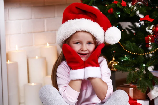Little girl in Santa hat and mittens sitting near fir tree on fireplace with candles background — Stok fotoğraf