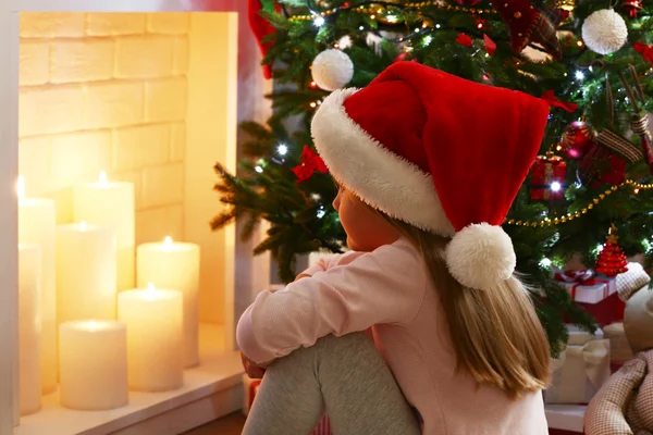 Niña en Santa sombrero sentado cerca de abeto en la chimenea con velas de fondo — Foto de Stock