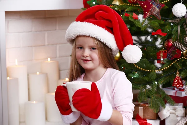 Bambina in cappello Babbo Natale e guanti prendere tazza seduta vicino abete sul camino con candele sfondo — Foto Stock