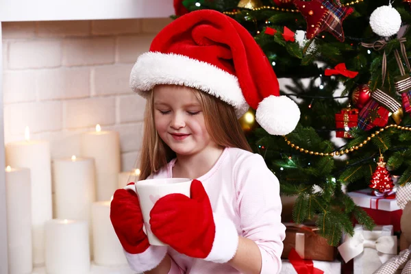 Petite fille dans le chapeau de Père Noël et mitaines prenant tasse assis près du sapin sur la cheminée avec fond de bougies — Photo