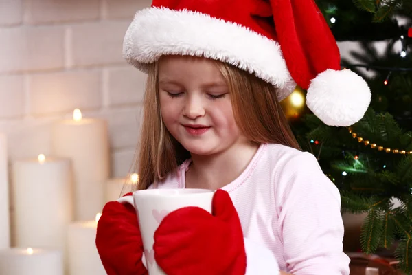Niña en Santa sombrero y manoplas tomando taza sentada cerca de abeto en la chimenea con velas de fondo —  Fotos de Stock