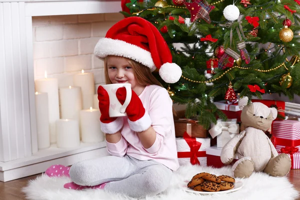 Little girl in Santa hat and mittens taking cup sitting near fir tree on fur carpet and wooden floor, on fireplace with candles background — 图库照片