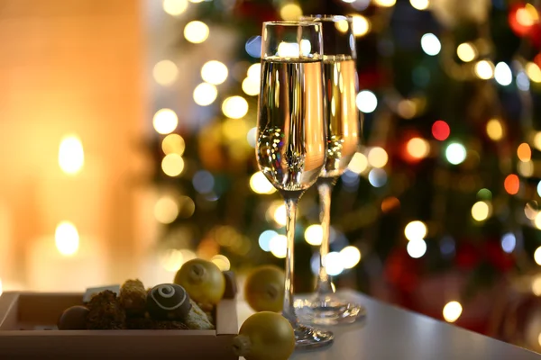 Two glass with champagne with chocolates and baubles on table on Christmas tree and fireplace background — Φωτογραφία Αρχείου