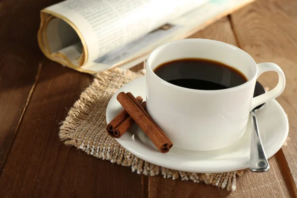 Xícara de café com pires, colher e canela em pano de serapilheira perto de jornal sobre fundo de mesa de madeira — Fotografia de Stock