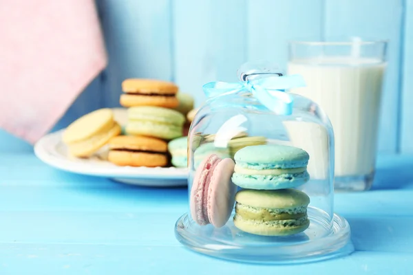 Gentle colorful macaroons in glass bell jar, milk glass and towel on color wooden table background — Stock Photo, Image