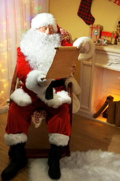 Санта-Клаус сидит со списком подарков в удобном кресле у камина дома — стоковое фото