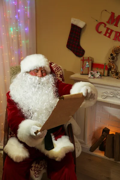 Santa Claus zitten met lijst van geschenken in comfortabele stoel in de buurt van open haard thuis — Stockfoto