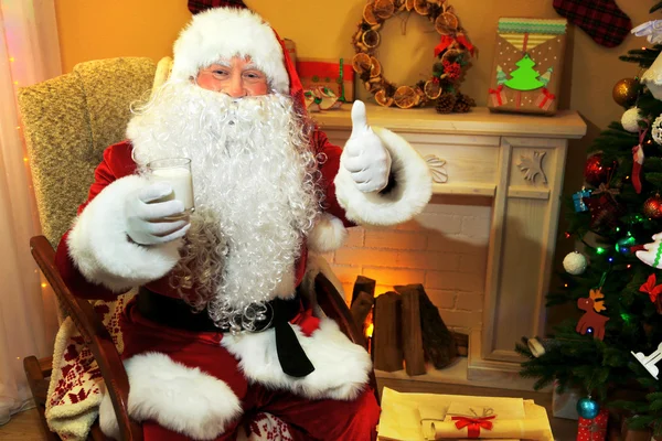Santa Claus zitten met melkglas in comfortabele stoel in de buurt van open haard thuis — Stockfoto