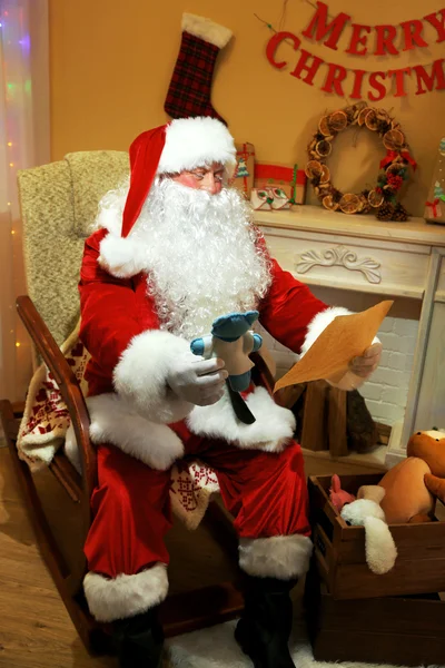 Santa Claus zitten met lijst van geschenken in comfortabele stoel in de buurt van open haard thuis — Stockfoto