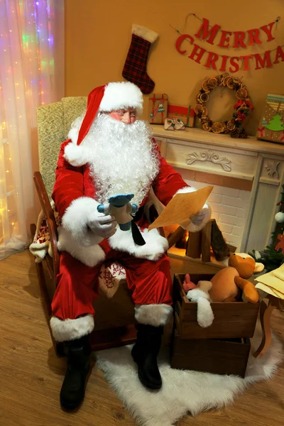 圣诞老人坐在舒适的椅子上，在家里壁炉旁的礼物列表 — 图库照片