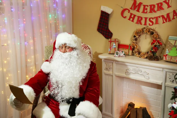 Santa Claus zitten met lijst van geschenken in comfortabele stoel in de buurt van open haard thuis — Stockfoto